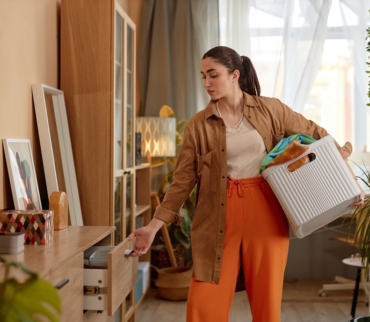 Mujer joven organizando un mueble del salón mientras sostiene una cesta con ropa, ejemplo de errores de organización comunes en espacios compartidos del hogar.