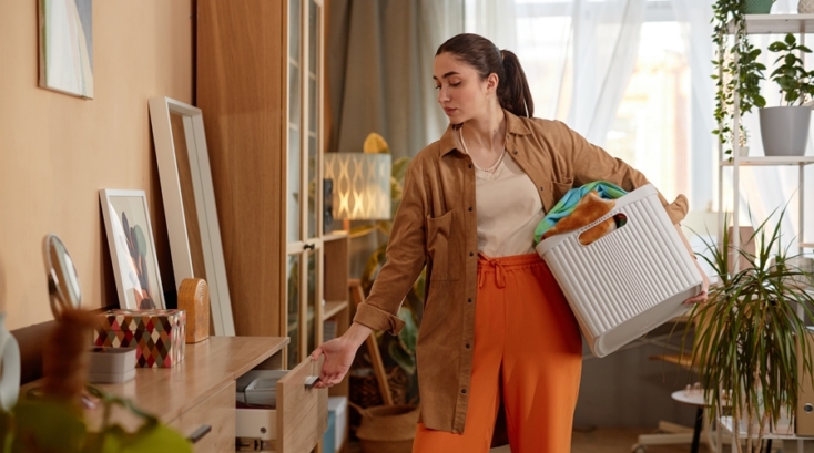 Mujer joven organizando un mueble del salón mientras sostiene una cesta con ropa, ejemplo de errores de organización comunes en espacios compartidos del hogar.