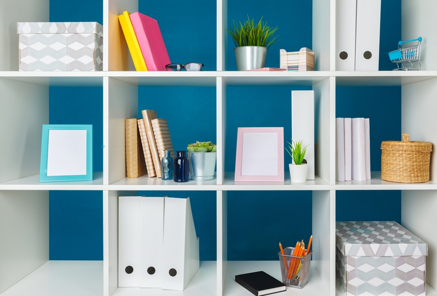 Estantería blanca con cajas, libros y plantas dispuestos de forma simétrica sobre fondo azul, un ejemplo visual de cómo el orden ayuda a reducir la ansiedad en entornos de trabajo o estudio.