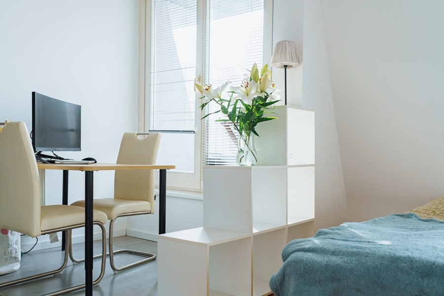 Zona de trabajo en vivienda pequeña con escritorio, ordenador y separador tipo estantería, como parte de un espacio multifuncional doméstico.