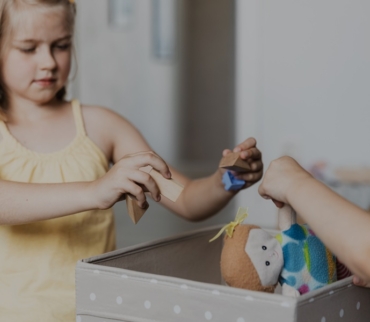 Niña colocando juguetes de madera y una muñeca en una caja gris, como parte de los hábitos de orden para niños.