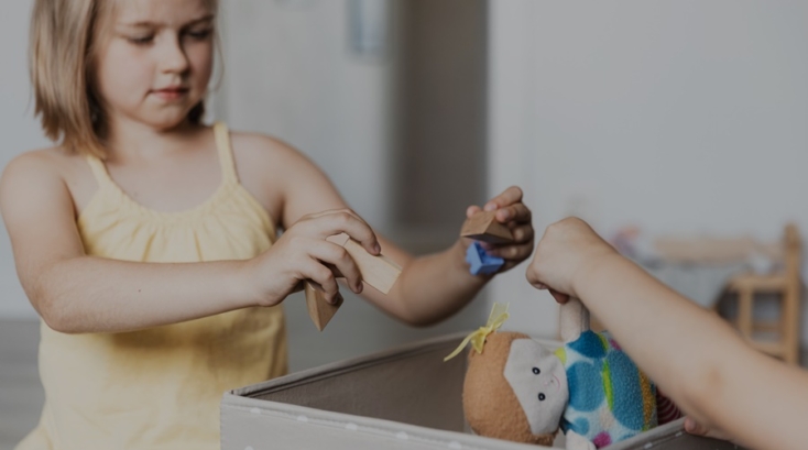 Niña colocando juguetes de madera y una muñeca en una caja gris, como parte de los hábitos de orden para niños.