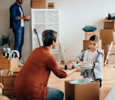 Mujer y niña empaquetando platos con papel protector mientras otra persona organiza cajas al fondo, en una casa llena de cajas de cartón, mostrando un proceso familiar y metódico para organizar mudanza de forma segura.
