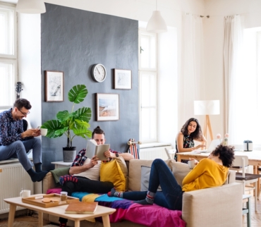 Cuatro compañeros de piso en un salón compartido, cada uno en diferentes actividades, en un ambiente luminoso y ordenado. Imagen ilustrativa de reglas de orden en un piso compartido.