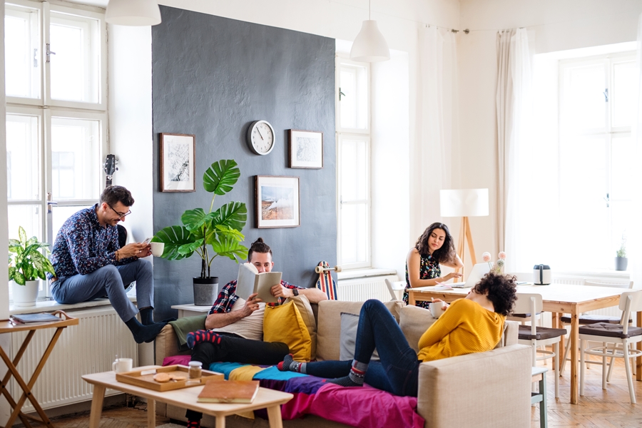 Cuatro compañeros de piso en un salón compartido, cada uno en diferentes actividades, en un ambiente luminoso y ordenado. Imagen ilustrativa de reglas de orden en un piso compartido.